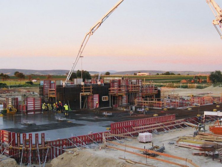 Photo: view of plant under construction, cranes, scaffolds, truck with mixing plant, access roads; landscape in the background, sky with evening light 
