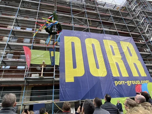 Blick auf eine Gebäuderohbaufassade, vor der ein Baugerüst steht. Ein gigantisches Baustellenbanner hängt am Gerüst mit dem Logo der Firma PORR. Auf dem Baugerüst steht ein Polier, über ihm schwebt der Richtkranz.