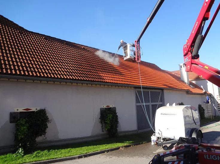 Foto: Dachbeschichtung: Von einem Krankorb aus wird das rote Ziegeldach von einem Arbeiter mit einem Hochruckwasserstrahlgerät gereinigt bevor die Beschichtungssubstanz auf das eingeschossige Haus aufgetragen werden kann.