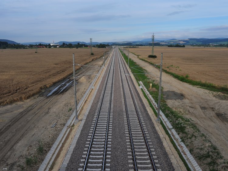 Photo: aerial view of two pairs of railway tracks through the fields in vanishing point perspective; power lines and catenary, muddy roads and fields on the side of the tracks, landscape in the background, cloudy evening sky 