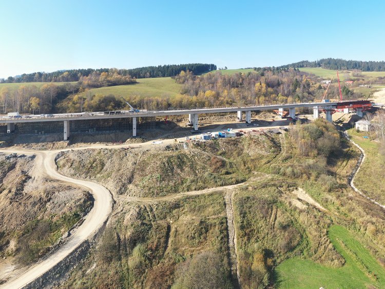 Photo: aerial view of a bridge under construction in rolling landscape; access roads in the foreground, forests and clear blue sky in the background